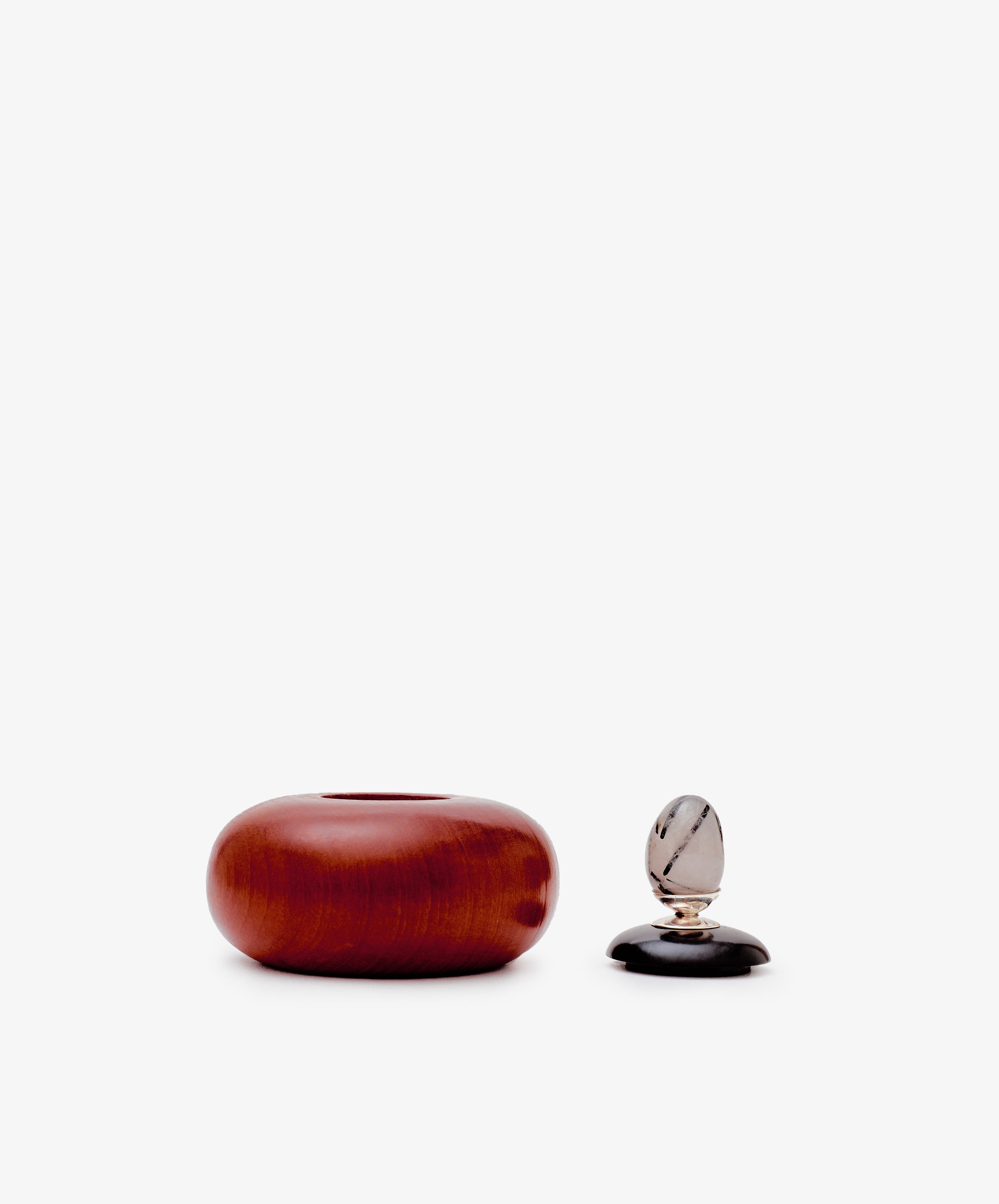 The Curiosity Box, a round reddish-brown African wood container, sits beside a small clear and black decorative item on a plain white background.