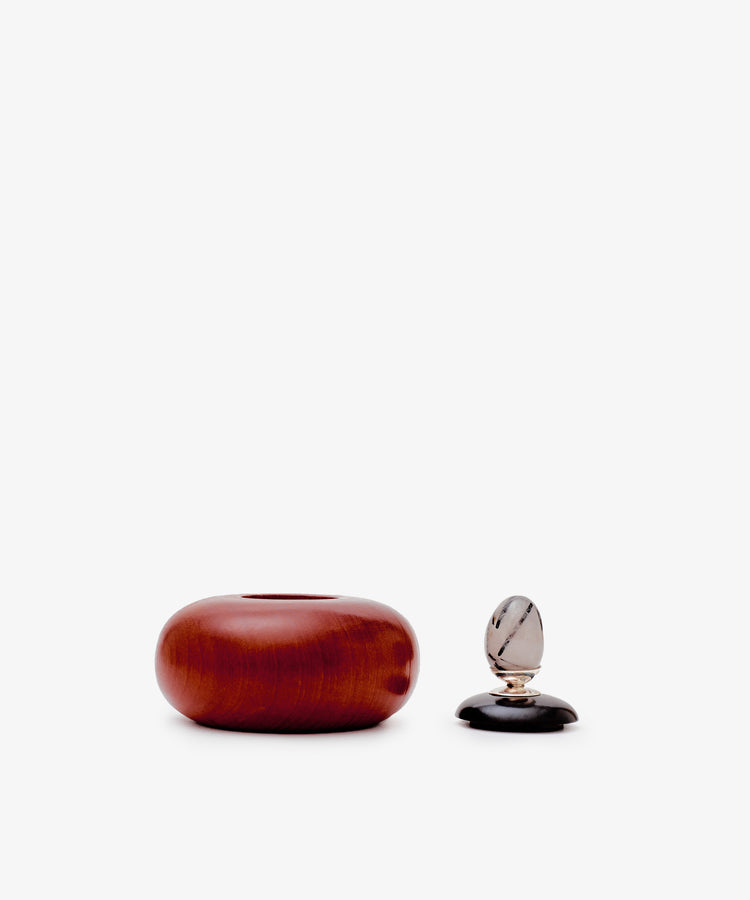The Curiosity Box, a round reddish-brown African wood container, sits beside a small clear and black decorative item on a plain white background.