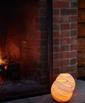 A lit fireplace with a black mesh screen stands beside two glowing, striped orange Akila Candleholders, handcrafted from Egyptian alabaster and resting on a stone hearth.