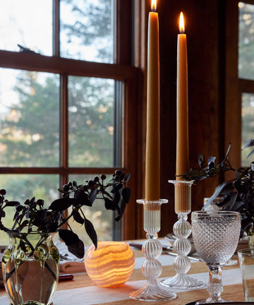A table by a window features two lit tall candles in Akila Candleholders of Egyptian Alabaster, a water glass, dark foliage in a vase, and a glowing round lamp.