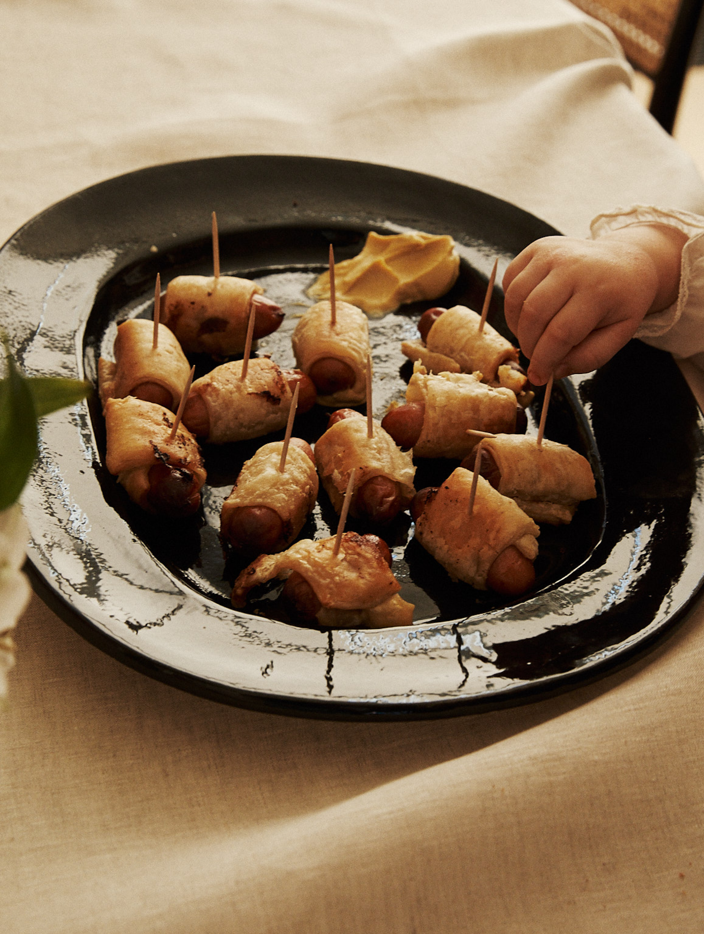 On the Small Rectangular Platter, pastry-wrapped mini sausages with toothpicks and mustard are served. A child's hand reaches to grab one of the savory snacks.