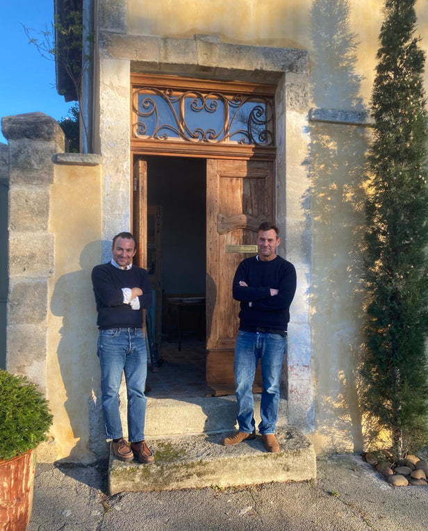 Two men in jeans and sweaters standing with arms crossed outside a rustic wooden doorway