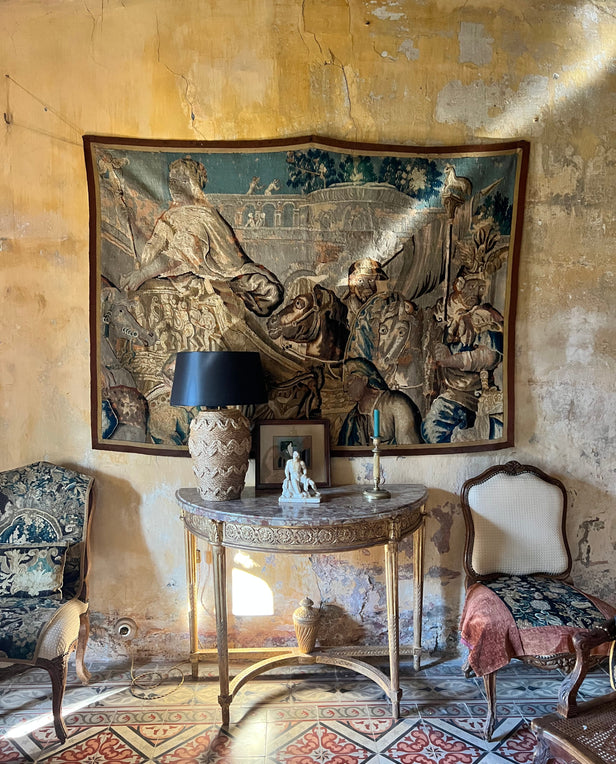 Antique room with a tapestry on the wall, ornate table with a lamp, and vintage chairs