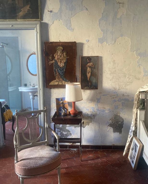 Vintage room with worn walls, a chair, side table with lamp, and religious and figure paintings