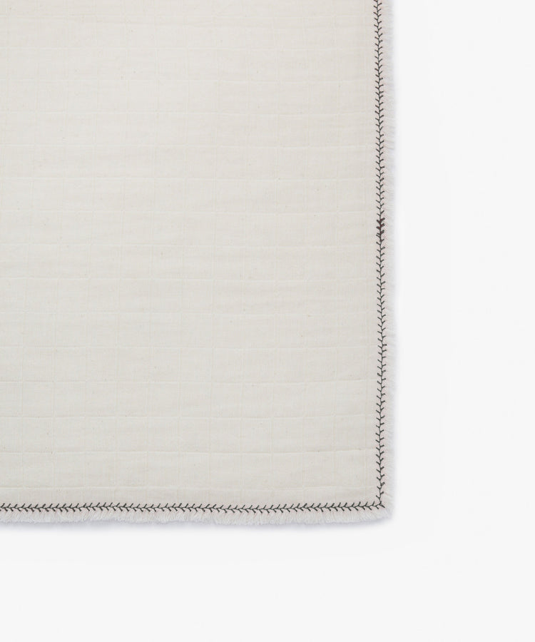 A close-up of the Filial Napkin, Set of 6, in cream with a grid pattern and dark branch stitching along the edge, displayed on a white tabletop background.