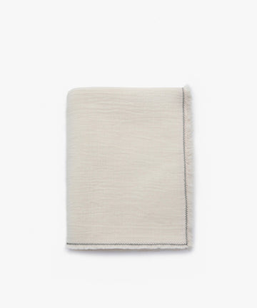 The Filial Tablecloth is a folded beige fabric with a visible zigzag stitch along the edge and natural weave texture, displayed on a plain white background.
