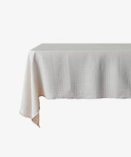 The Filial Tablecloth, featuring a white natural weave and subtle grid pattern, drapes elegantly over the corner of a rectangular table for a minimal look against a plain white background.
