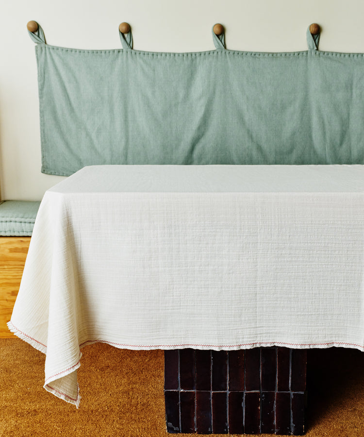 A Filial Tablecloth in white natural weave covers a table placed before a cushioned bench with a green fabric backrest against the wall.