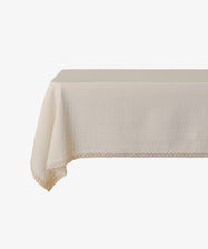 The Double Gauze Flower Tablecloth, in rectangular beige cotton with subtle texture and decorative lace trim, is draped over a table against a white background.