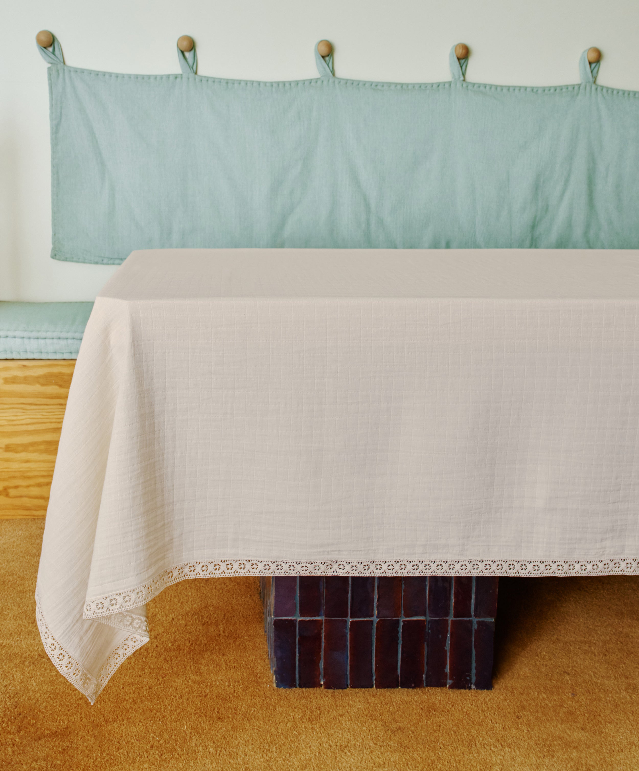 The Double Gauze Flower Tablecloth, featuring a delicate lace trim, drapes softly over a rectangular table set before a cushioned bench with light green upholstery.