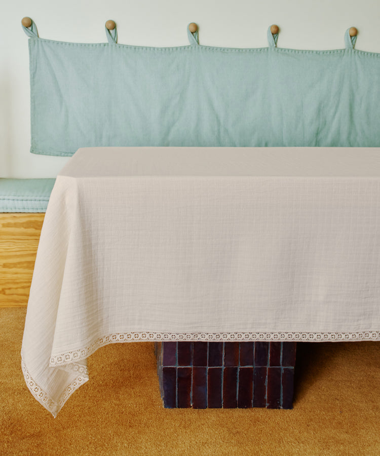 The Double Gauze Flower Tablecloth, featuring a delicate lace trim, drapes softly over a rectangular table set before a cushioned bench with light green upholstery.