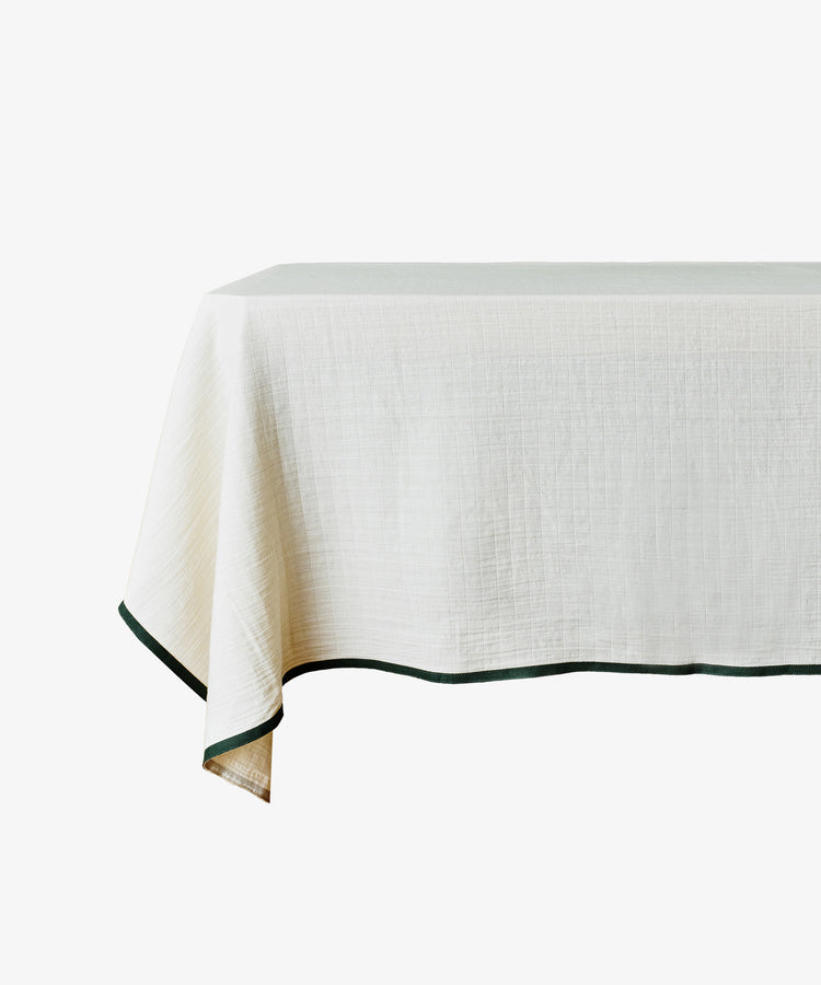 The Fita Tablecloth is a rectangular, light-colored cotton tablecloth with a dark border along the edge, offering an organic texture.