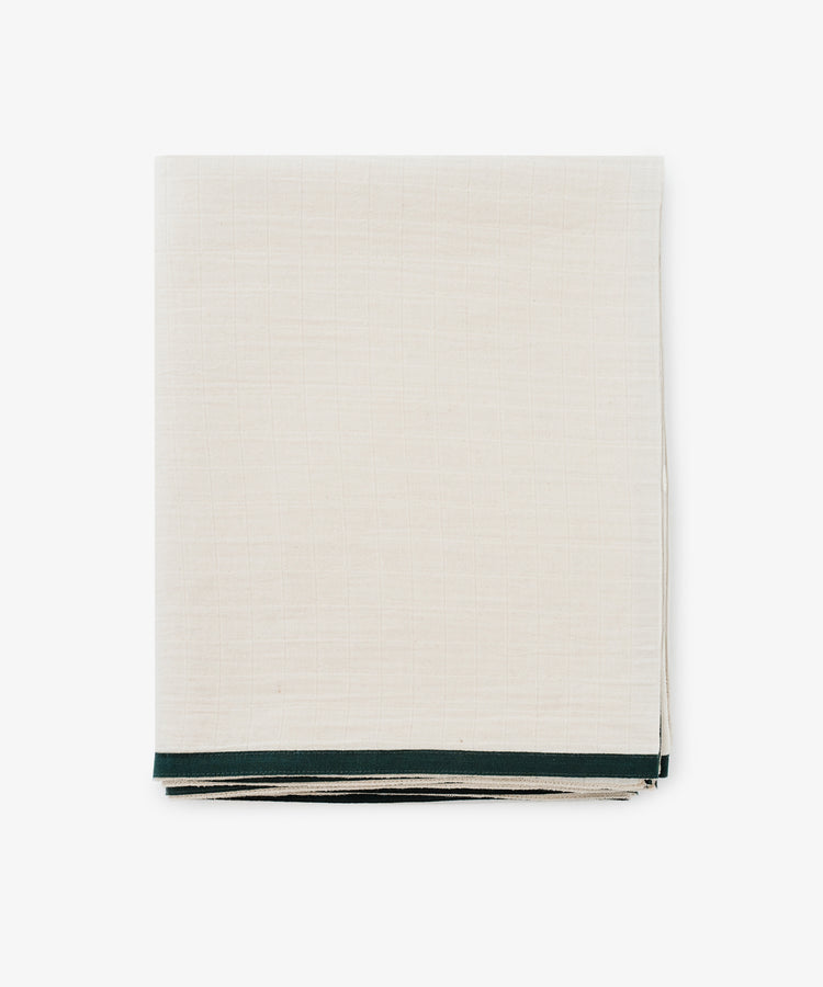 The Fita Tablecloth, made from natural cotton, features a neatly folded off-white fabric with a thin dark green border and is displayed on a plain white background.