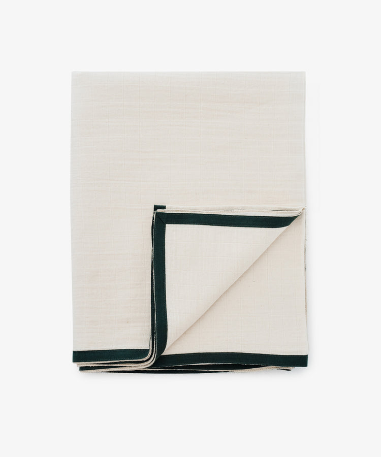 The Fita Tablecloth, made from natural cotton, features a folded beige design with a dark green border and is displayed flat on a white surface to highlight its organic texture.