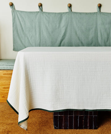A rectangular table with a Fita Tablecloth in natural cotton white sits in front of a built-in bench with green cushions on a tan carpeted floor.