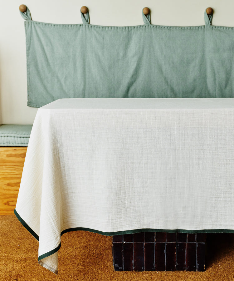 A rectangular table with a Fita Tablecloth in natural cotton white sits in front of a built-in bench with green cushions on a tan carpeted floor.