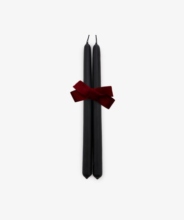 A set of two Tapered Candles in matte black are elegantly bound with a dark red ribbon and displayed against a plain white background. Hand-dipped for quality, these candles combine sophisticated style with a long burn time, making them ideal for any occasion.