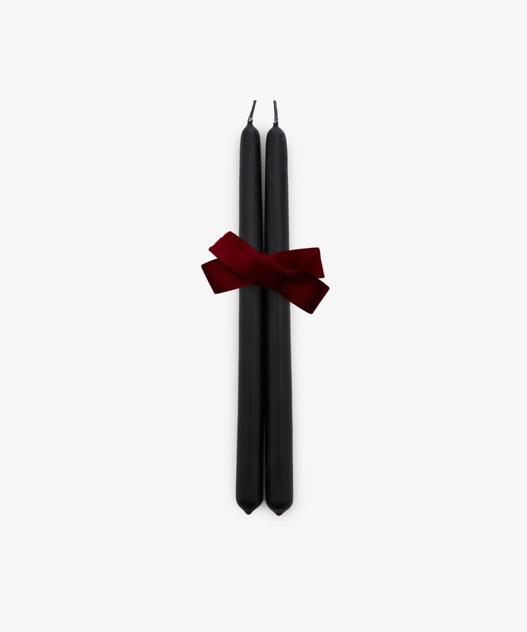 A set of two Tapered Candles in matte black are elegantly bound with a dark red ribbon and displayed against a plain white background. Hand-dipped for quality, these candles combine sophisticated style with a long burn time, making them ideal for any occasion.