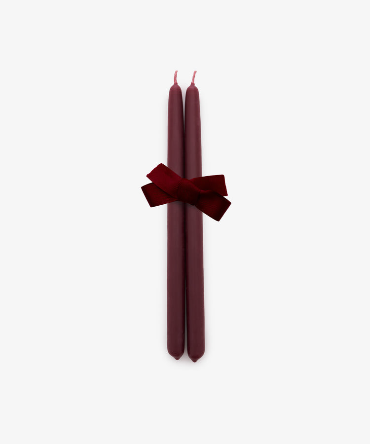 A set of two berry red Tapered Candles, hand-dipped and tied together with a matching red ribbon bow, is displayed against a plain white background. Their classic design and extended burn time make them an elegant choice for any occasion.