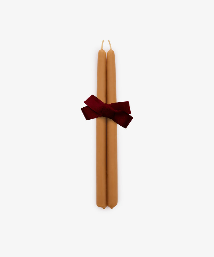 The Tapered Candles, Set of 2, features two hand-dipped tan taper candles tied together with a dark red ribbon and displayed against a plain white background.