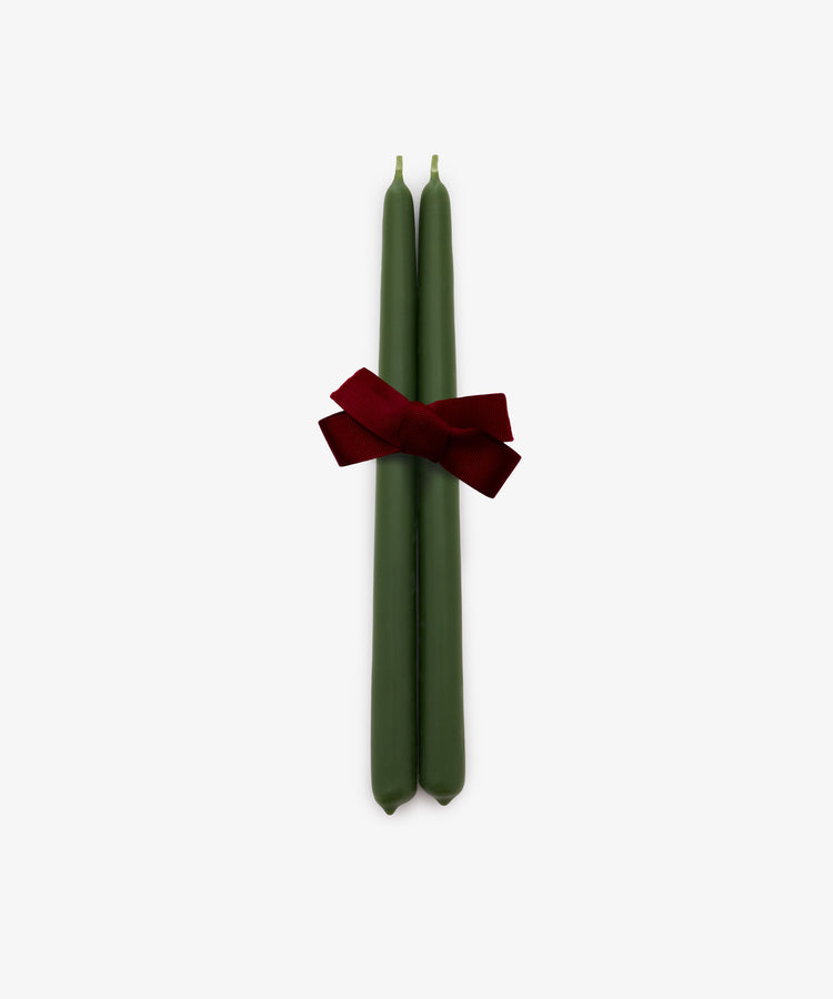 A set of two hand-dipped, mossy green Tapered Candles are tied together with a red ribbon bow and displayed against a plain white background. These elegant candles provide a long burn time, making them ideal for creating a warm and inviting atmosphere.