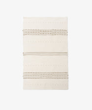 The Gaia Floor Mat is a rectangular cream-colored rug featuring subtle raised dots and horizontal brown stripes, inspired by Portuguese tradition and shown on a white background.