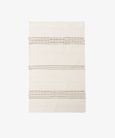 The Gaia Floor Mat is a rectangular cream-colored rug featuring subtle raised dots and horizontal brown stripes, inspired by Portuguese tradition and shown on a white background.