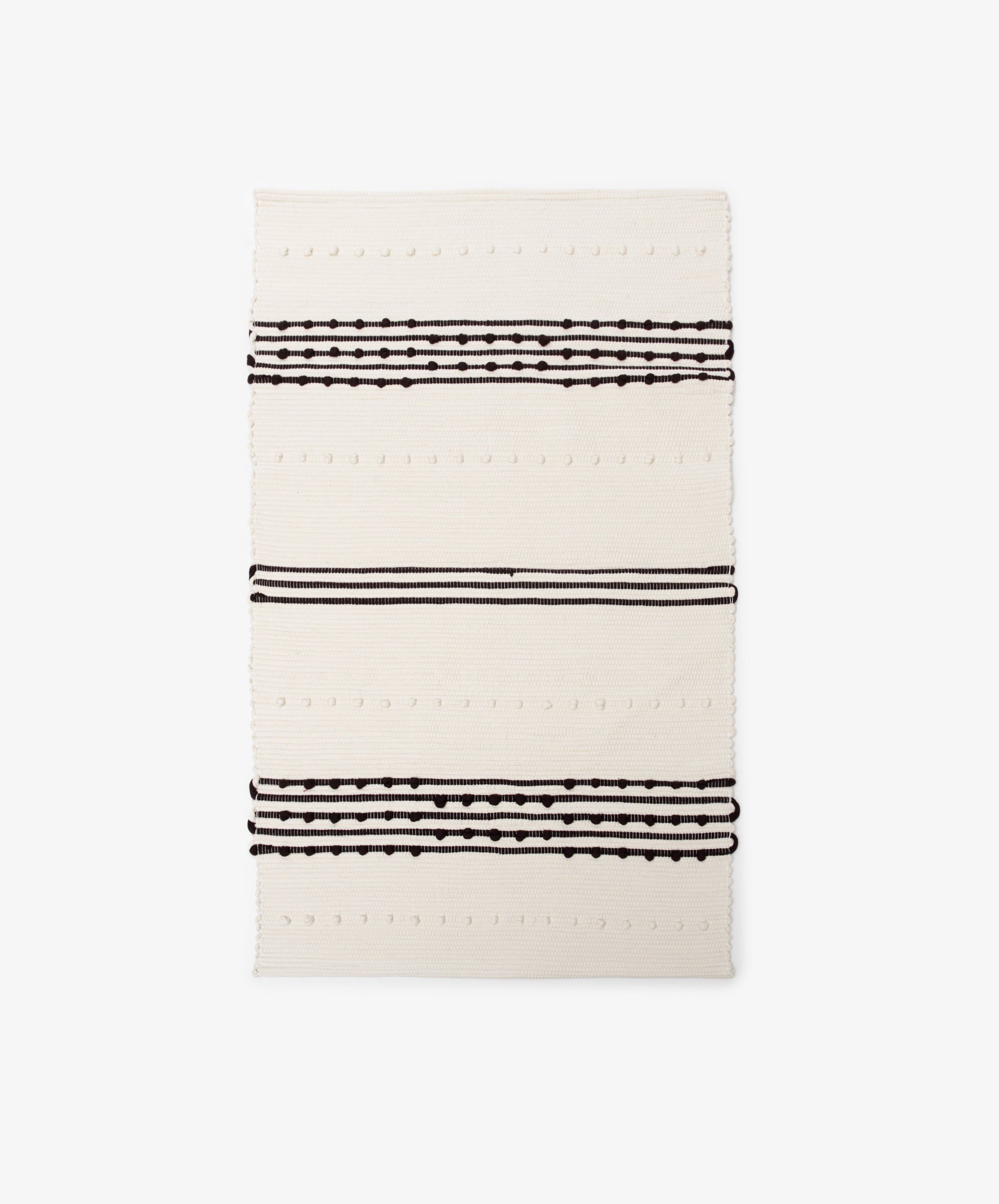The Gaia Floor Mat is a rectangular cream-colored rug inspired by Portuguese tradition, featuring three sets of parallel black lines and dotted patterns across its width.