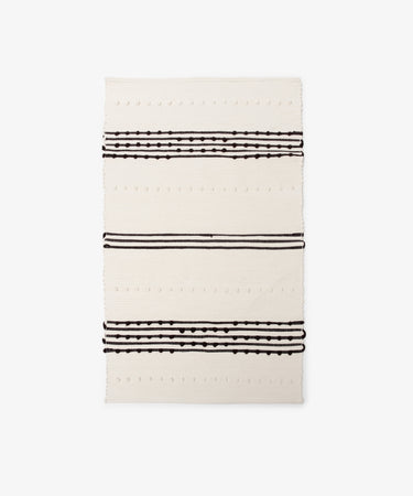 The Gaia Floor Mat is a rectangular cream-colored rug inspired by Portuguese tradition, featuring three sets of parallel black lines and dotted patterns across its width.