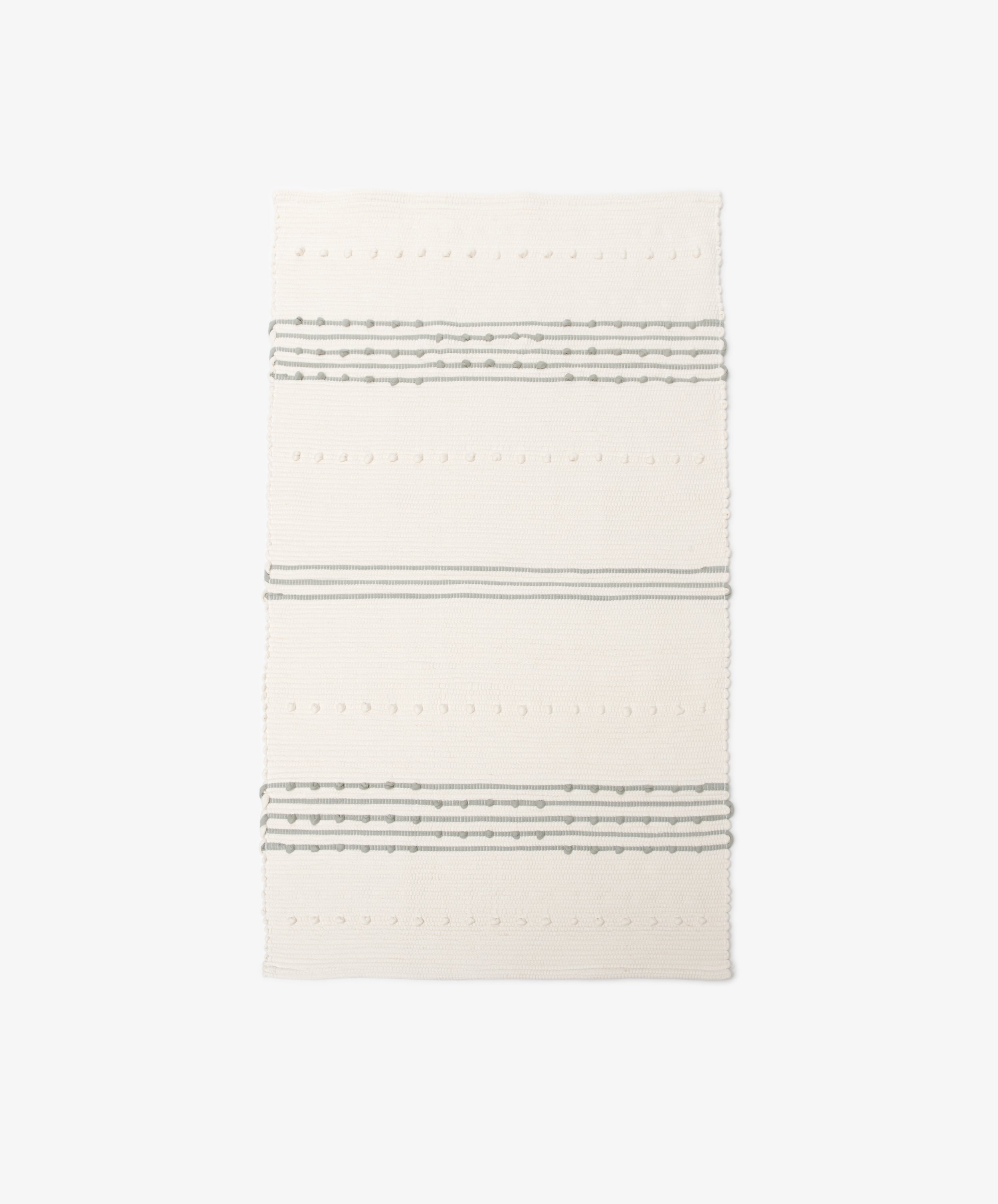 The Gaia Floor Mat is a rectangular, cream-colored rug handwoven in the Portuguese tradition from recycled fabric, with three sets of parallel gray stripes and textured dot patterns on a plain white background.