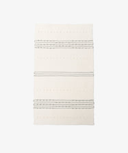 The Gaia Floor Mat is a rectangular, cream-colored rug handwoven in the Portuguese tradition from recycled fabric, with three sets of parallel gray stripes and textured dot patterns on a plain white background.