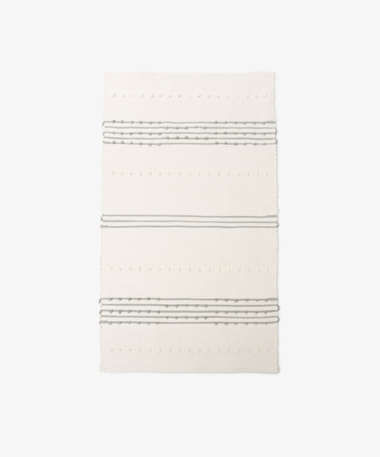The Gaia Floor Mat is a rectangular, cream-colored rug handwoven in the Portuguese tradition from recycled fabric, with three sets of parallel gray stripes and textured dot patterns on a plain white background.