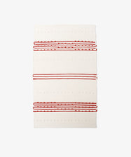 The Gaia Floor Mat is a rectangular, cream-colored rug inspired by Portuguese tradition, made from recycled fabric with textured red and white stripes and raised dot patterns.