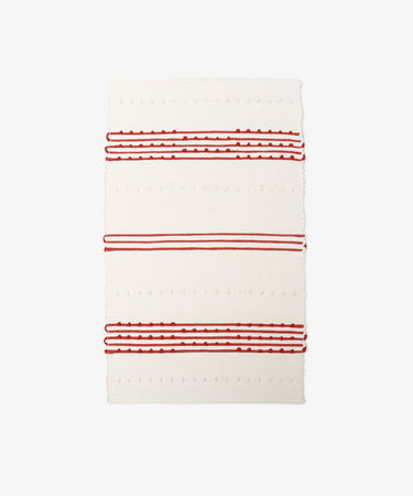 The Gaia Floor Mat is a rectangular, cream-colored rug inspired by Portuguese tradition, made from recycled fabric with textured red and white stripes and raised dot patterns.