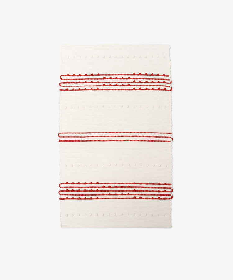The Gaia Floor Mat is a rectangular, cream-colored rug inspired by Portuguese tradition, made from recycled fabric with textured red and white stripes and raised dot patterns.