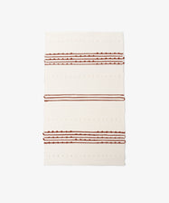 The Gaia Floor Mat is a rectangular cream-colored rug inspired by Portuguese tradition, handwoven from recycled fabric with horizontal raised dots and three sets of brown and rust stripes for an eco-friendly accent.