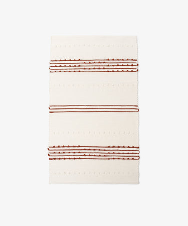 The Gaia Floor Mat is a rectangular cream-colored rug inspired by Portuguese tradition, handwoven from recycled fabric with horizontal raised dots and three sets of brown and rust stripes for an eco-friendly accent.