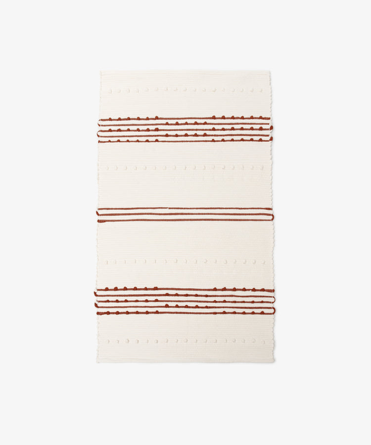 The Gaia Floor Mat is a rectangular cream-colored rug inspired by Portuguese tradition, handwoven from recycled fabric with horizontal raised dots and three sets of brown and rust stripes for an eco-friendly accent.