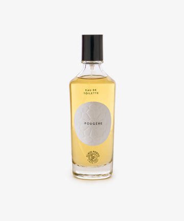 A clear glass bottle with light amber liquid, black cap, and a white label reading Fougère Eau de Toilette, evoking pine needle and cedarwood notes.
