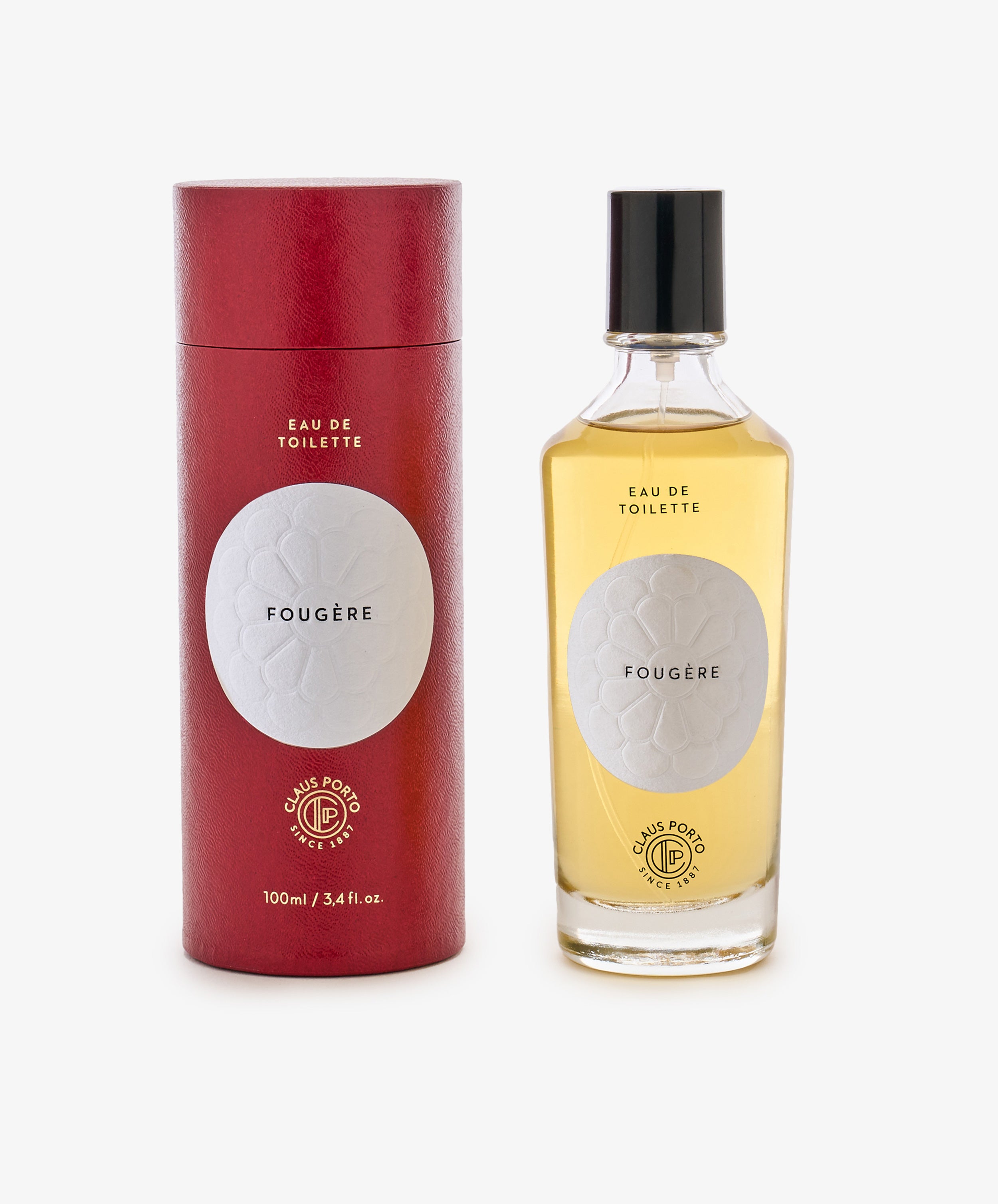 A bottle of Fougère Eau de Toilette sits beside its red cylindrical package, both with white labels and black text, evoking pine needle and cedarwood notes.