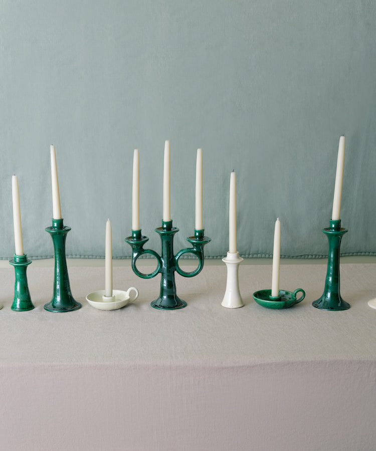 Grouping of various green and white ceramic candleholders with white taper candles lined up on grey tablecloth.