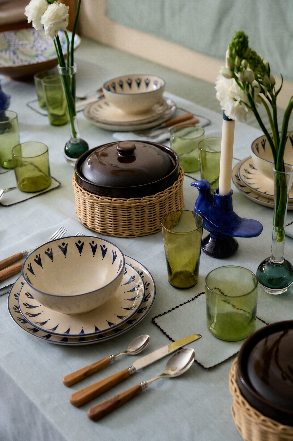 A dining table set with patterned plates, green glasses, wooden cutlery, ceramic bowls with lids, vases with flowers, and a blue bird-shaped candleholder rests atop the Nico Tablecloth in a soft mint blue.