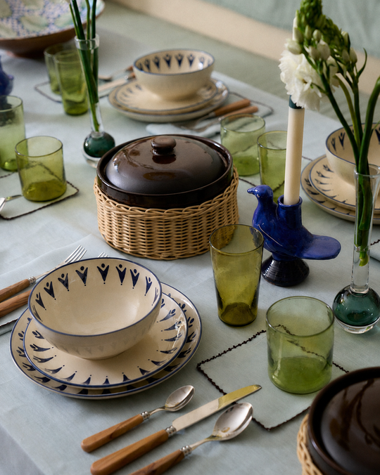 A dining table set with patterned plates, green glasses, wooden cutlery, ceramic bowls with lids, vases with flowers, and a blue bird-shaped candleholder rests atop the Nico Tablecloth in a soft mint blue.