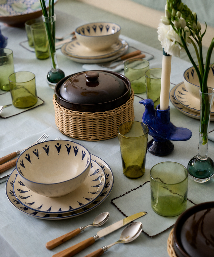 A dining table set with patterned plates, green glasses, wooden cutlery, ceramic bowls with lids, vases with flowers, and a blue bird-shaped candleholder rests atop the Nico Tablecloth in a soft mint blue.
