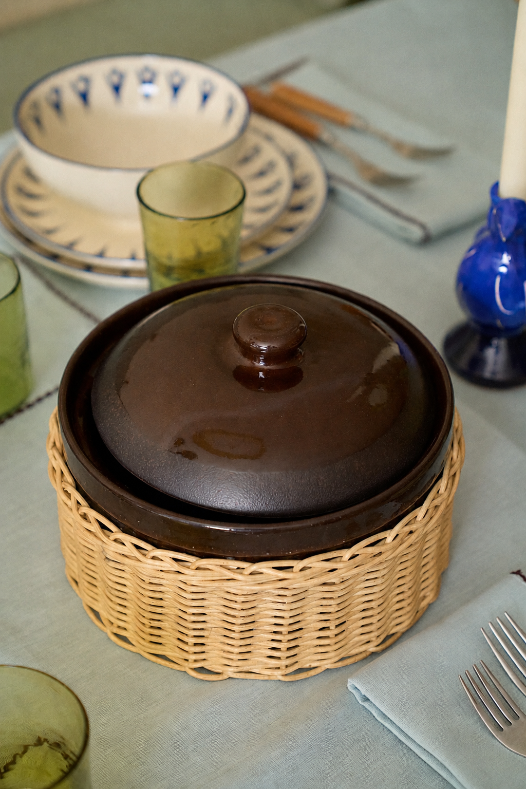An Amany Casserole in brown ceramic with a lid rests in a woven cover on a set dining table surrounded by plates, utensils, glasses, and a blue candle.