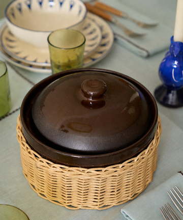 An Amany Casserole in brown ceramic with a lid rests in a woven cover on a set dining table surrounded by plates, utensils, glasses, and a blue candle.