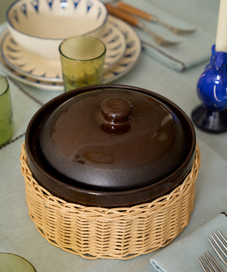 An Amany Casserole in brown ceramic with a lid rests in a woven cover on a set dining table surrounded by plates, utensils, glasses, and a blue candle.