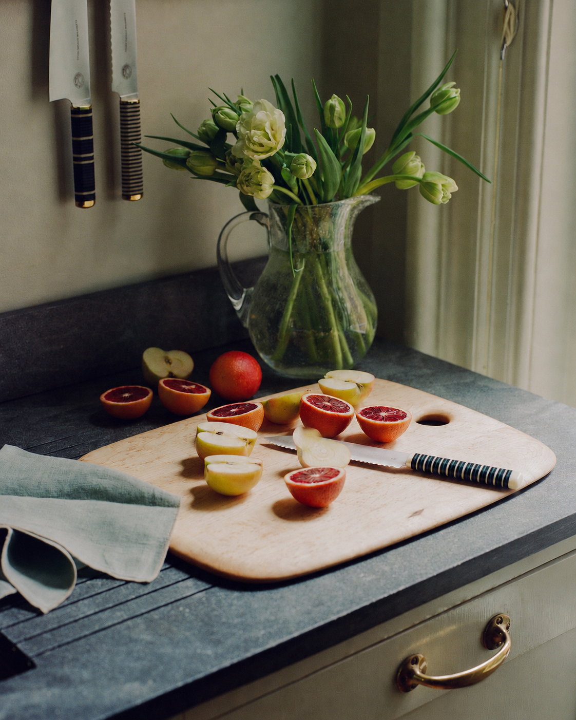 A cutting board with halved plums and the Bread Knife, which features a forest green handle, sits on a kitchen counter near a pitcher of flowers under a window.