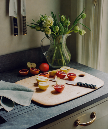 A cutting board with halved plums and the Bread Knife, which features a forest green handle, sits on a kitchen counter near a pitcher of flowers under a window.