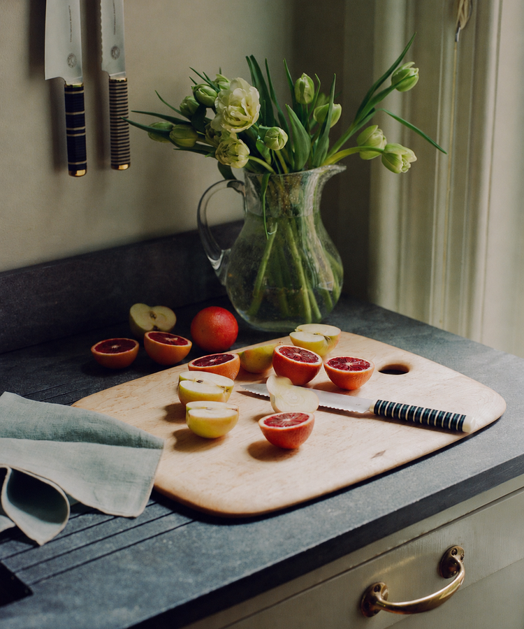A cutting board with halved plums and the Bread Knife, which features a forest green handle, sits on a kitchen counter near a pitcher of flowers under a window.